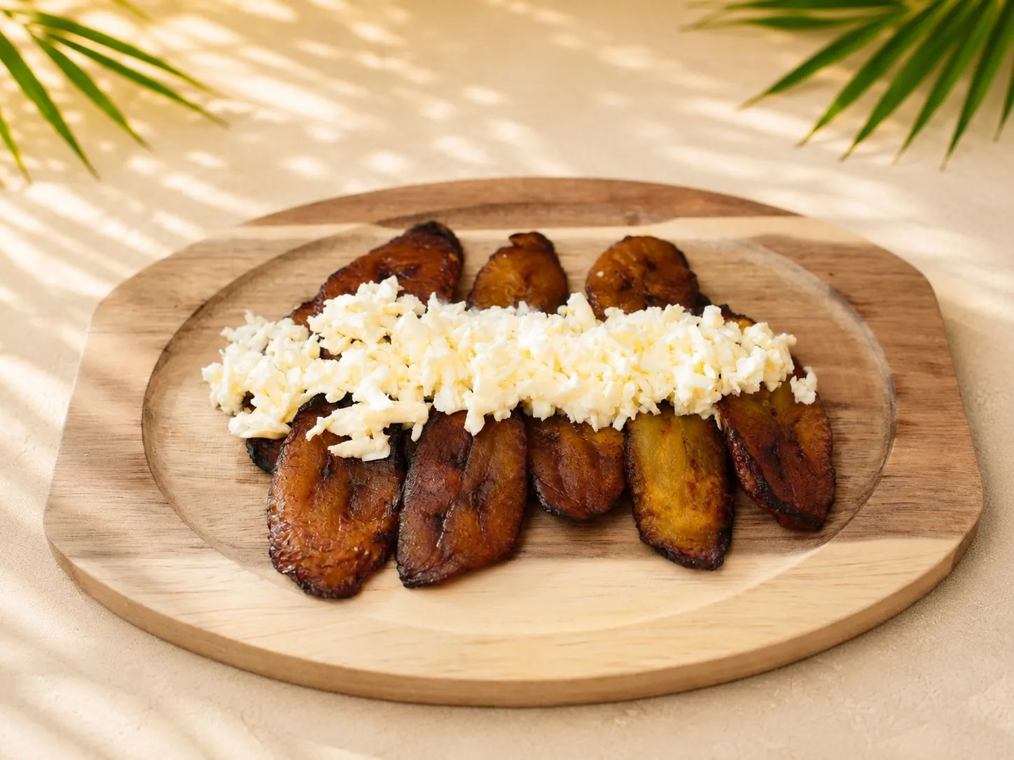 Tajadas: gebratene reife Kochbananenscheiben mit zerbröseltem Hirtenkäse auf einem Holzbrett.
